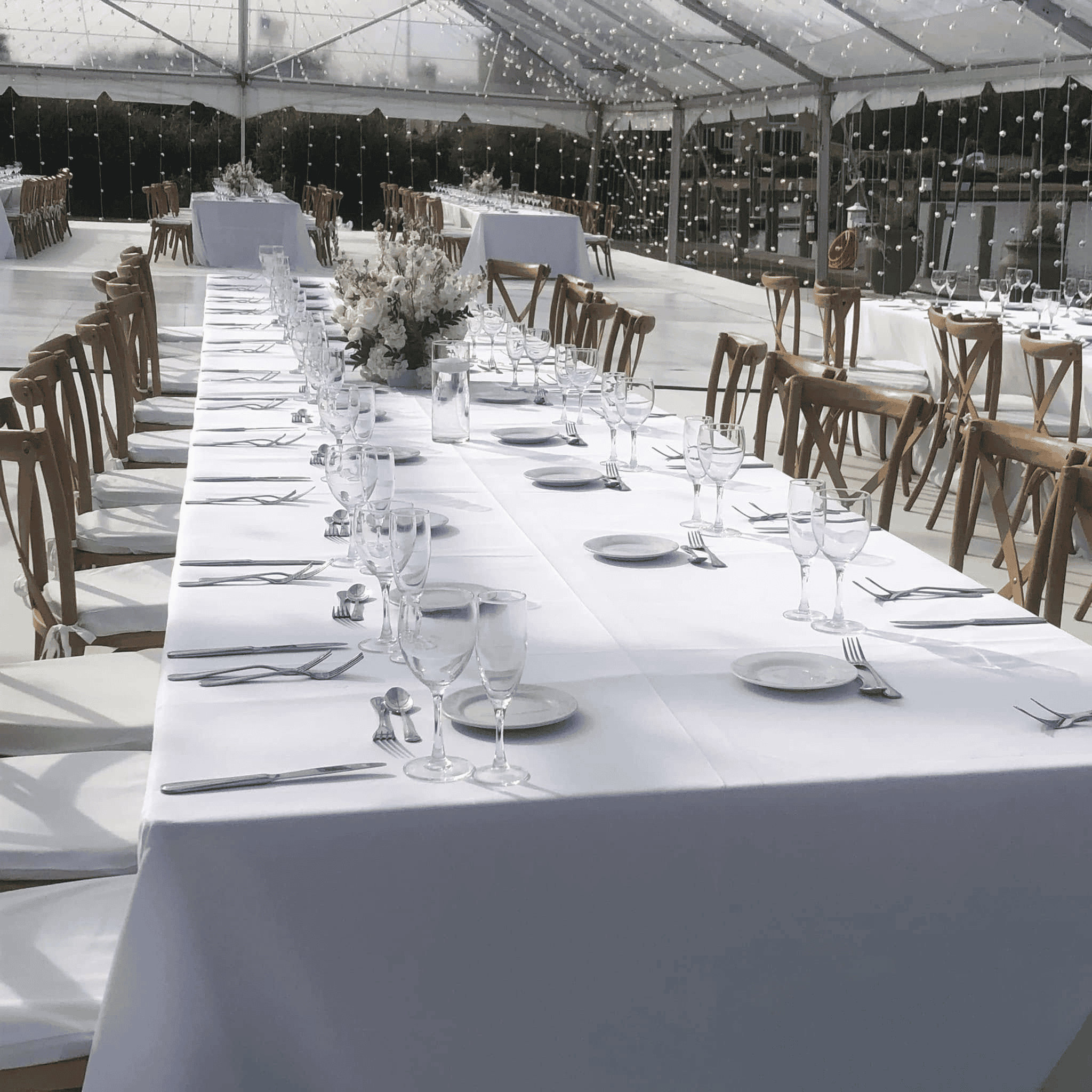 Long banquet table set with glassware and wooden cross back chairs under a clear top tent