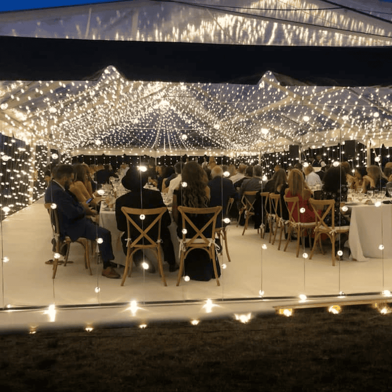Reception under a canopy of fairy lights with guests at wooden tables on a white floor