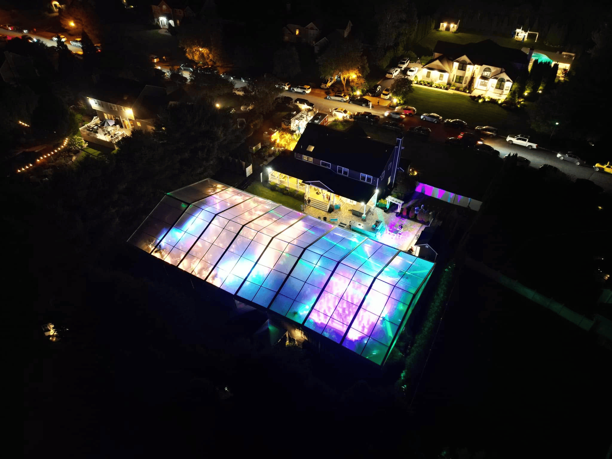 Aerial view at night of a long clear top tent lit in purple and green beside a house