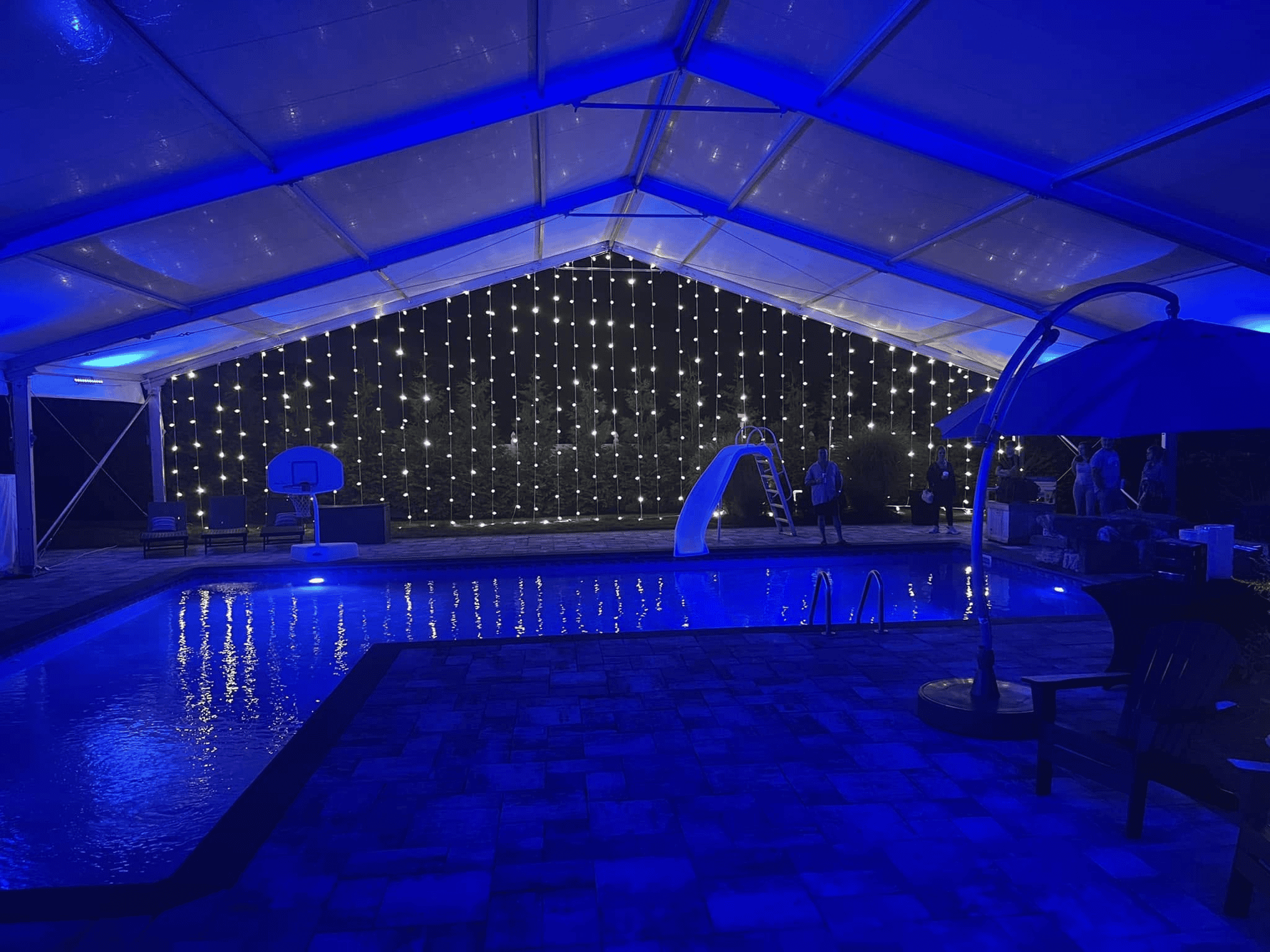 Clearspan tent beside a swimming pool at night with blue uplighting and a curtain of string lights