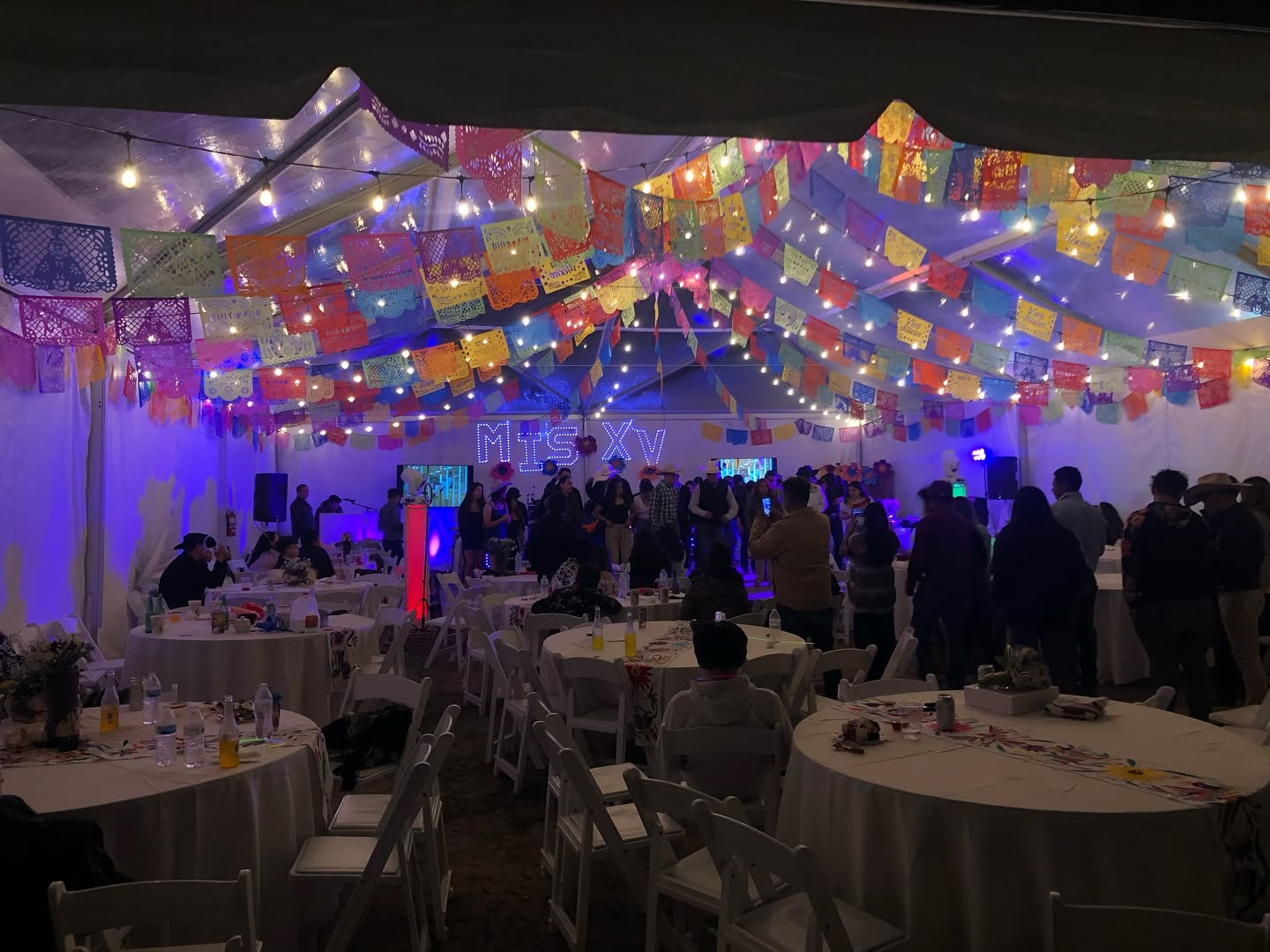 Quinceanera under a tent at night with papel picado banners, string lights and guests at round tables