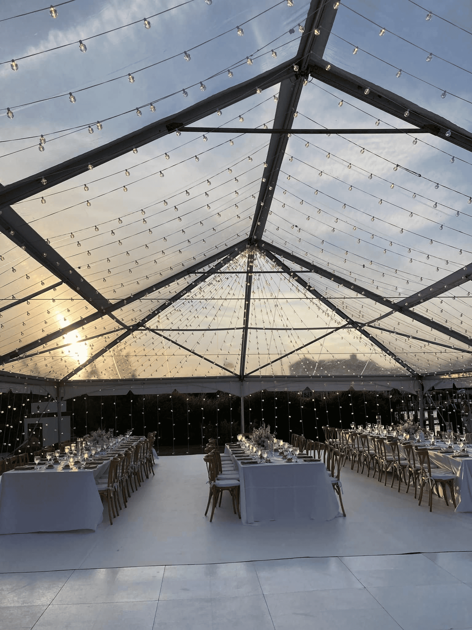 Clear top tent at sunset with string lights across the roof and long tables set with chargers and glassware